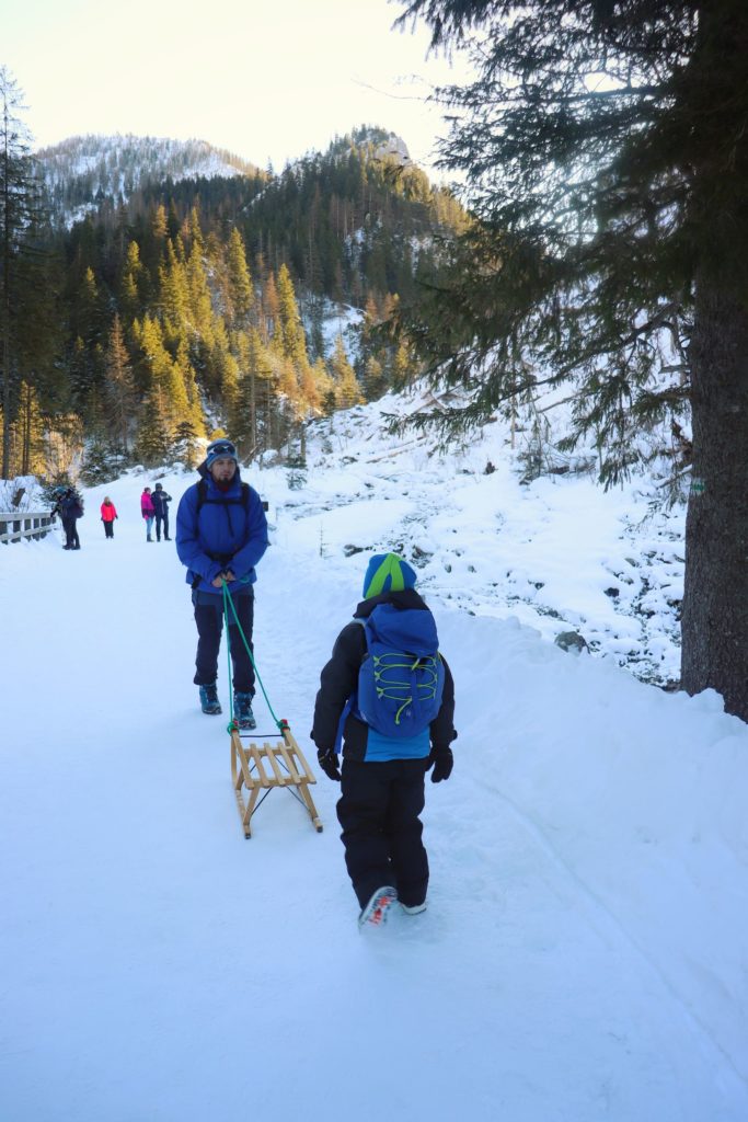 dwóch turystów, ojciec ciągnący sanki, dziecko idące pieszo, w tle tatrzańskie szczyty oraz most