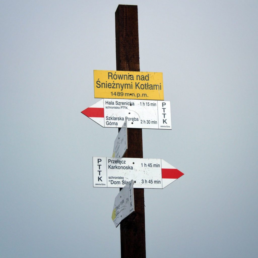 Żółta tablica z napisem Równia nad Śnieżnymi Kotłami leżąca 1489 metrów nad poziomem morza, inne tabliczki opisujące idące stąd szlaki w kolorze czerwonym