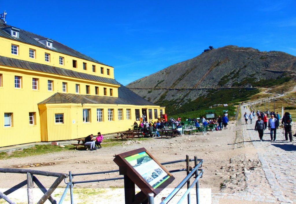 Żółty budynek - Dom Śląski leżący u podnóża szczytu Śnieżka, sporo turystów, w oddali widoczny szczyt Śnieżki