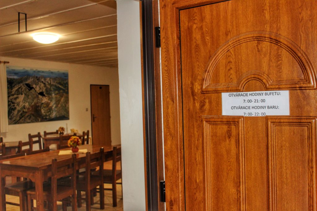 Drewniane drzwi prowadzące do Bufetu Rohackiego, na drzwiach kartka godzinami otwarcia bufetu, po lewej stronie widoczne wnętrze bufetu - stoliki z krzesłami