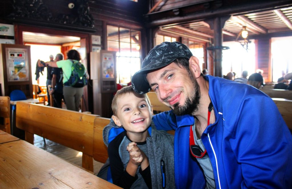 Dwóch turystów, Tata oraz szczęśliwe dziecko w Domu Śląskim, widoczne drewniane wnętrze schroniska, w tle inni turyści