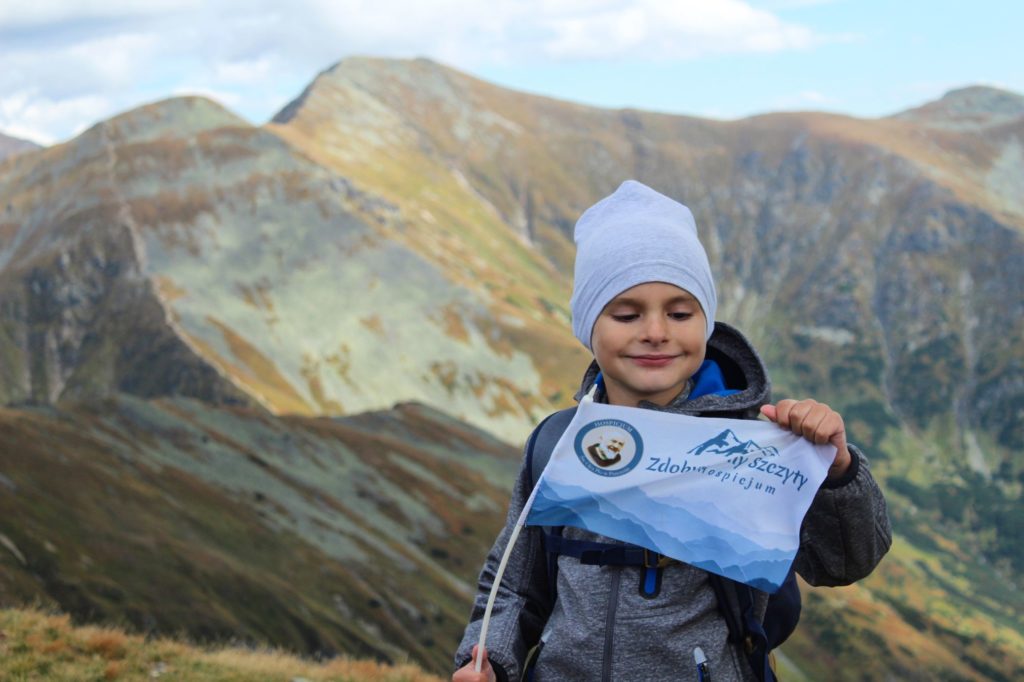 Szczęśliwe dziecko na Wołowcu trzymające horongiewkę z napisem Zdobywamy Szczyty dla Hospicjum na tle Tatr