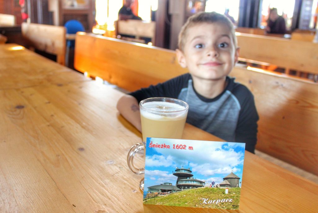 Uśmiechnięte dziecko w Domu Śląskim siedzące przy drewnianym stole, na którym leży kawa oraz widokówka ze szczytu Śnieżka 1602 metry nad poziomem morza