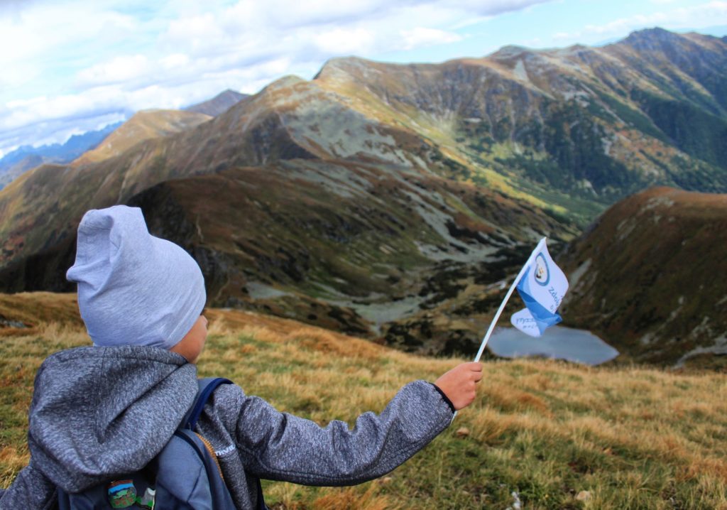 Wołowiec z dzieckiem, dziecko macha flagą akcji zdobywamy szczyty dla hospicjum, w tle tatrzańskie szczyty oraz staw leżący między górami