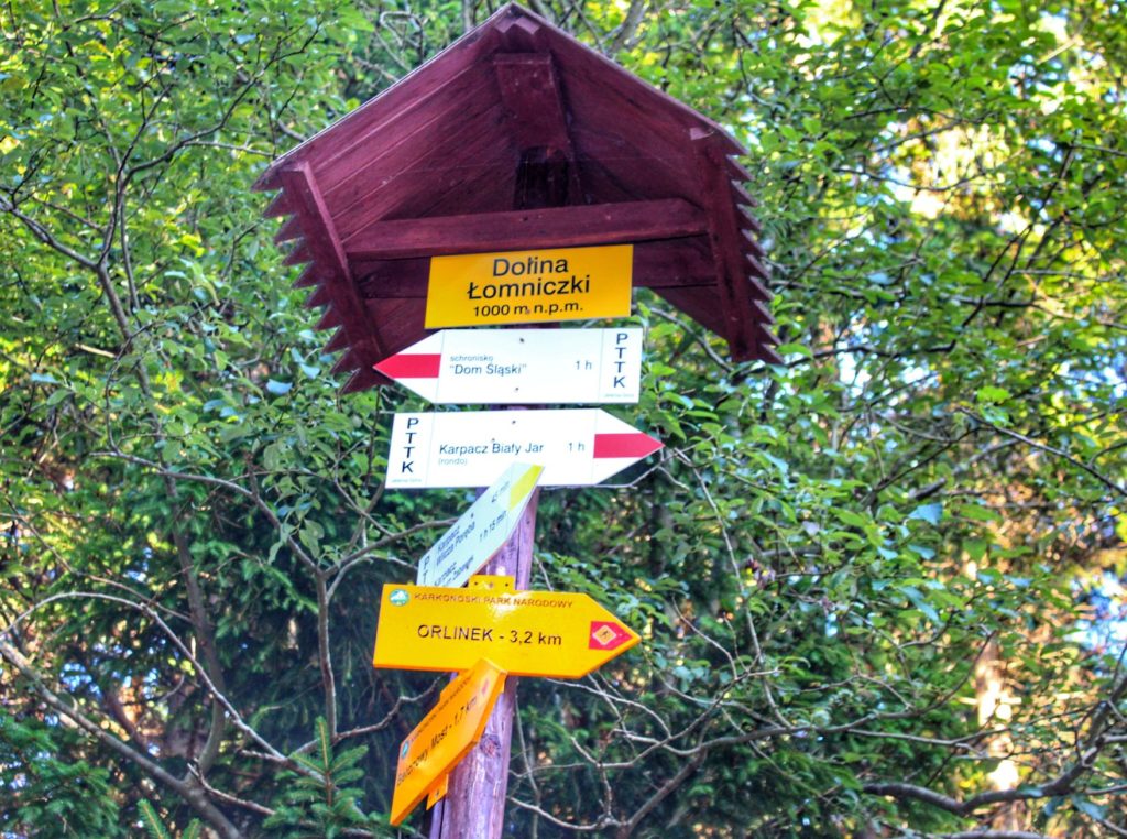 Żółta tabliczka z napisem Dolina Łomniczki leżąca 1000 metrów nad poziomem morza, inne tabliczki opisujące szlaki czerwone
