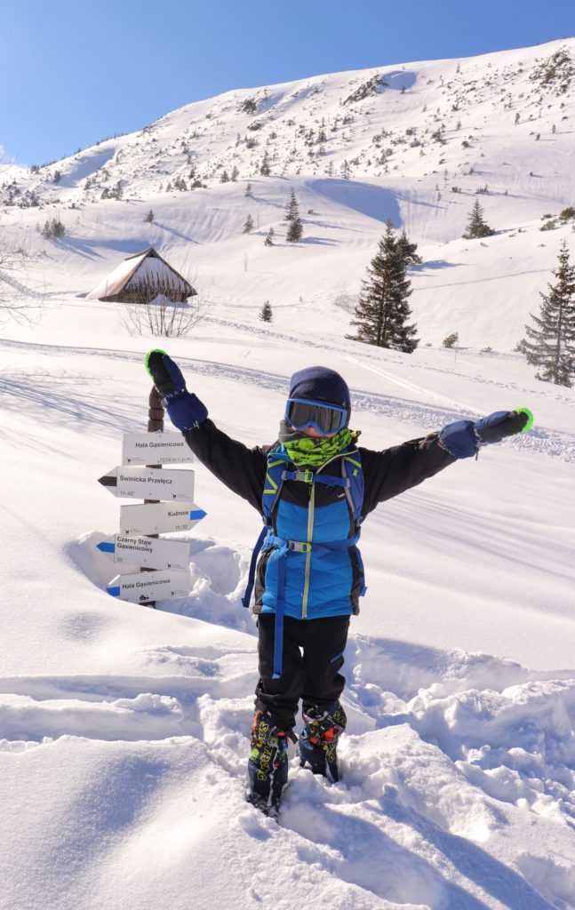 Hala Gąsienicowa z dzieckiem, szczęśliwe dziecko pozujące przy zasypanym przez śnieg słupie z białymi tabliczkami oznaczającymi Halę Gąsienicową, w tle zabytkowa chatka, błękitne niebo
