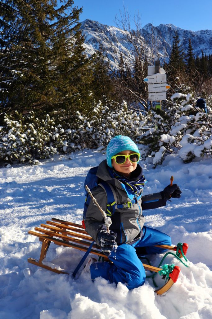 Zadowolone dziecko siedzące na sankach przy słupie z tabliczką na Hali Gąsienicowej, zimowe Tatry