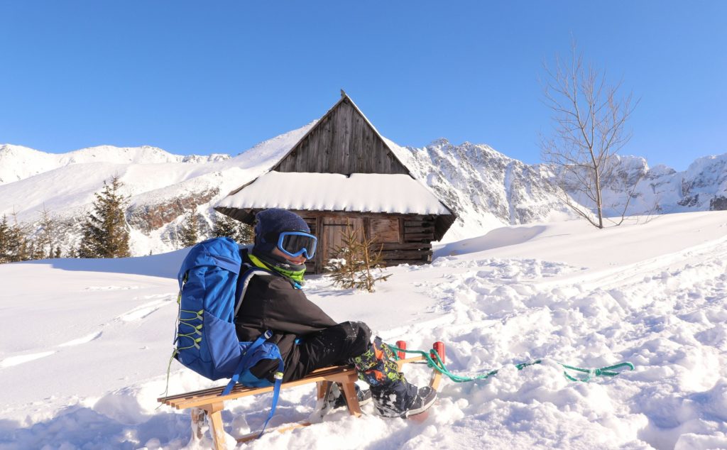 zadowolone dziecko siedzące na sankach na Hali Gąsienicowej zimą, w tle zabytkowa chata, piękny, słoneczny dzień