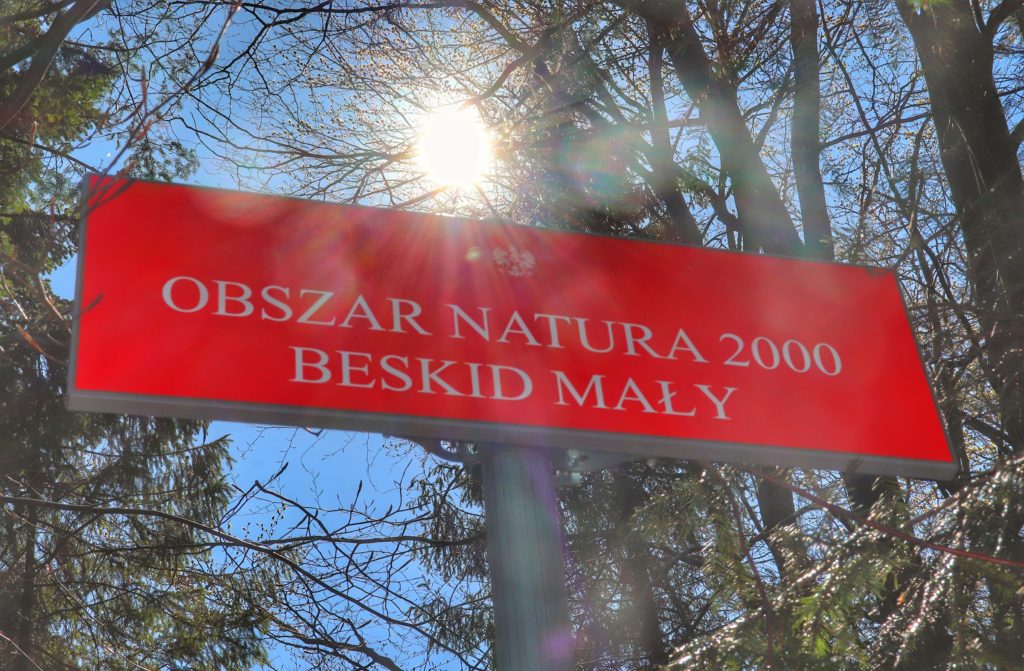 Czerwona tablica z napisem OBSZAR NATURA 2000 BESKID MAŁY w okolicach Przysłopu, tablica oświetlona przez słońce