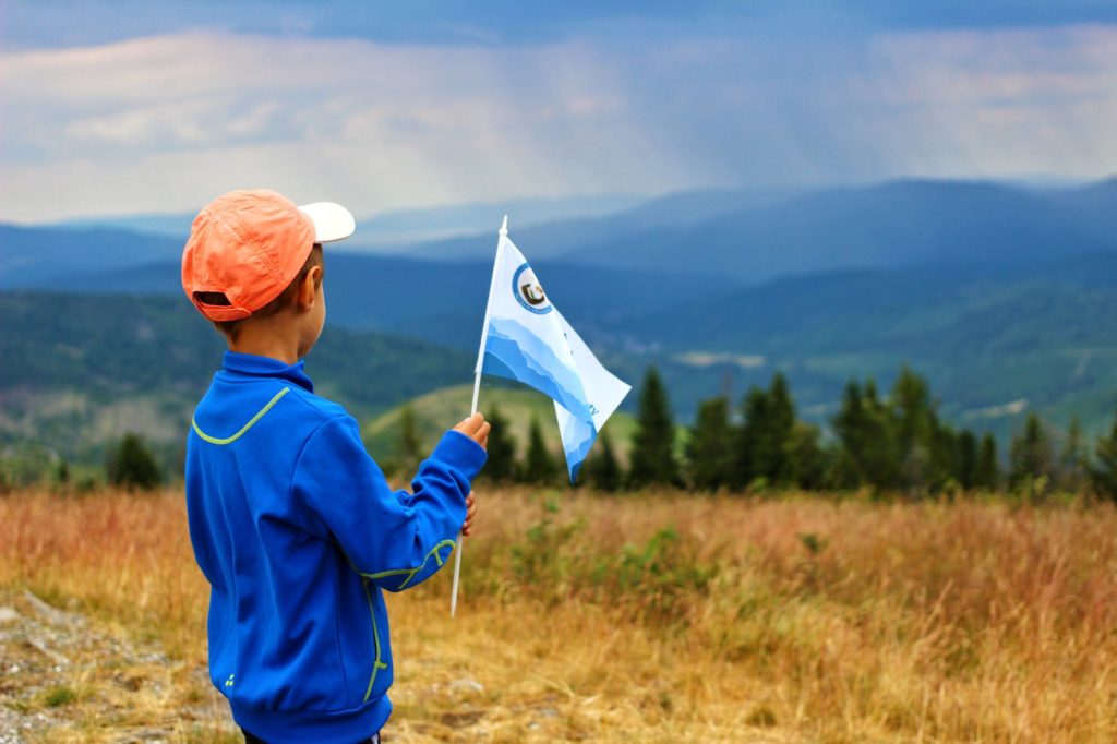 Dziecko na szczycie Bendoszka Wielka, trzymające w rączkach flagę akcji Zdobywamy Szczyty dla Hospicjum, w oddali widok na Beskid Żywiecki