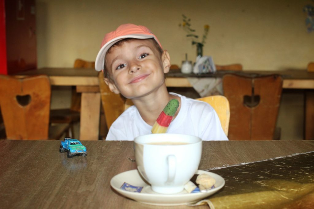Przegibek z dzieckiem, uśmiechnięty chłopiec siedzący w schronisku na Przegibku, jedzący loda,na drewnianym stole leży biała filiżanka z kawą oraz niebieski resorak