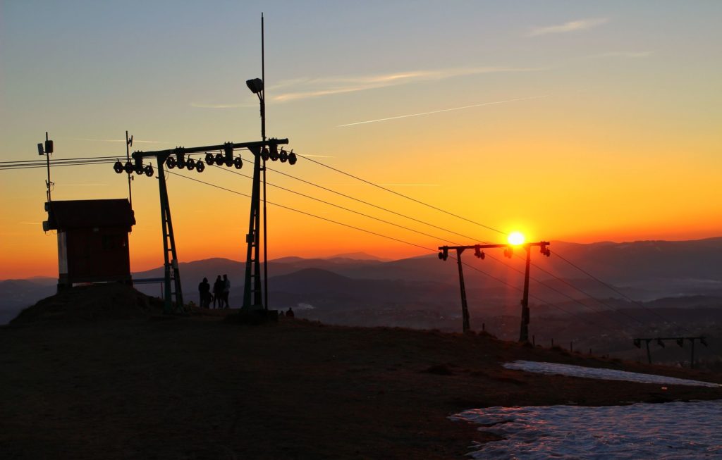 Wyciąg narciarski na szczycie Ochodzita oświetlony przez zachodzące za góry słońce, kolorowe niebo, grupka turystów, pozostałości śniegu