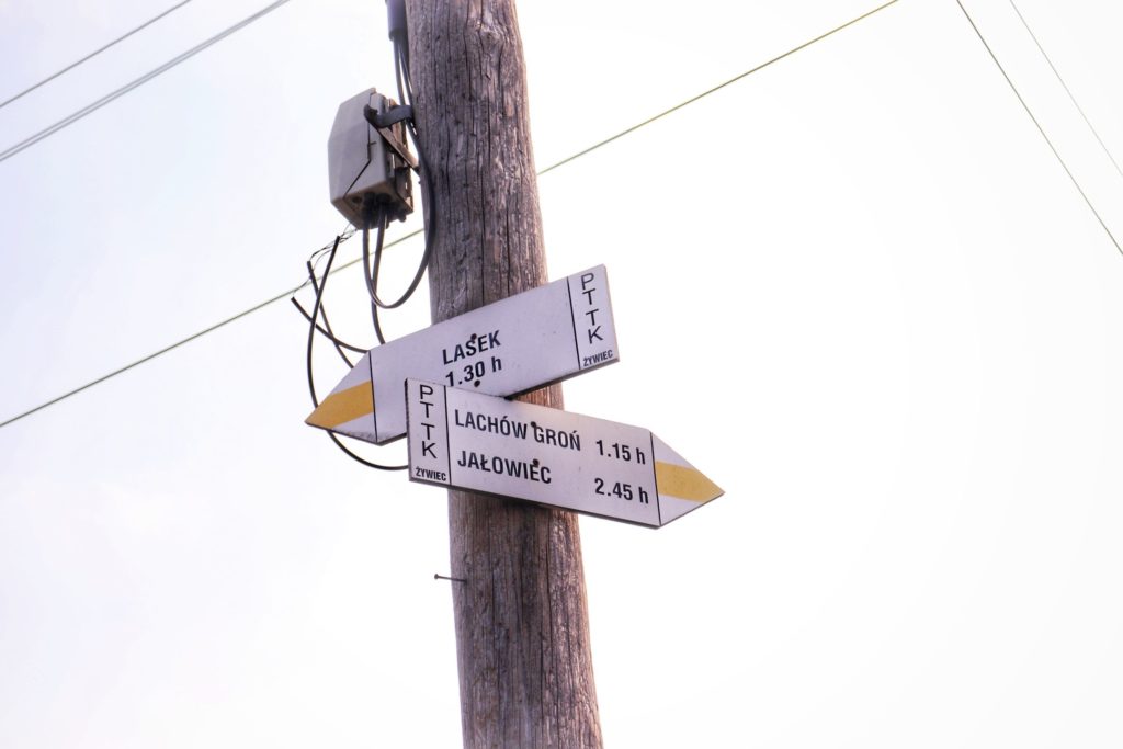 Dwie tablice, drogowskazy wiszące na drewnianym słupie w Koszarawie, opisujące żółty szlak prowadzący w dwóch kierunkach. Jedna z tablic z napisem LASEK czas przejścia 1.15 h, druga tablica z napisem Lachów Groń czas 1.15 h oraz Jałowiec 2.45 h
