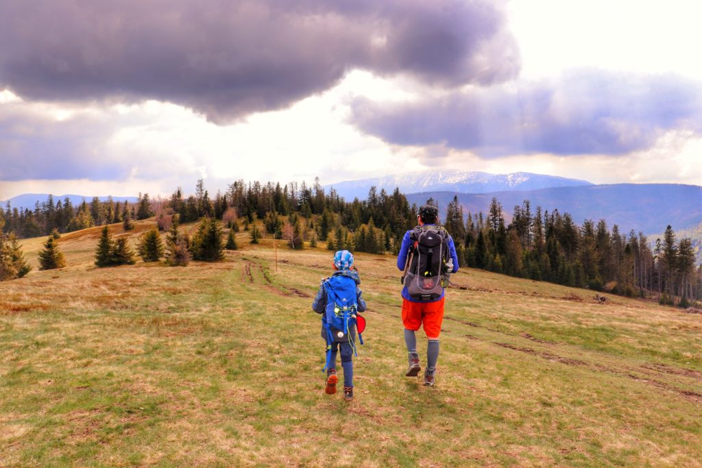 Dwóch turystów, ojciec z dzieckiem idący wzdłuż Hali Janoszkowej, zachmurzone niebo, w oddali widoczna ośnieżona Babia Góra