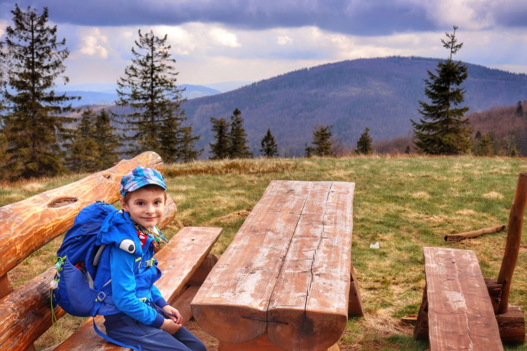 Szczyt Lachów Groń, Zadowolone dziecko siedzące przy drewnianym stole na ławce. W oddali widoczny szczyt Jałowiec, zachmurzone niebo