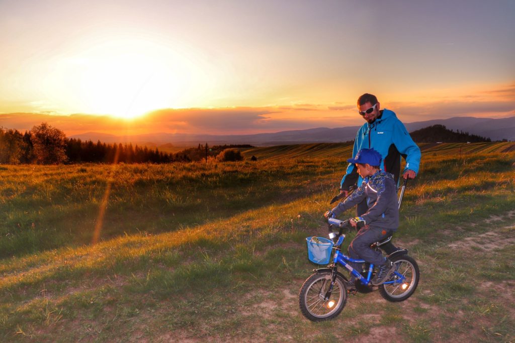 Turysta z dzieckiem na rowerku na tle zachodzącego słońca, w oddali krajobraz górski