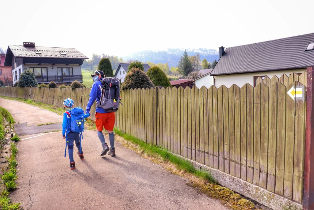 Turyści, mężczyzna z sześcioletnim dzieckiem idący żółtym szlakiem z Koszarawy, droga asfaltowa prowadząca między domami