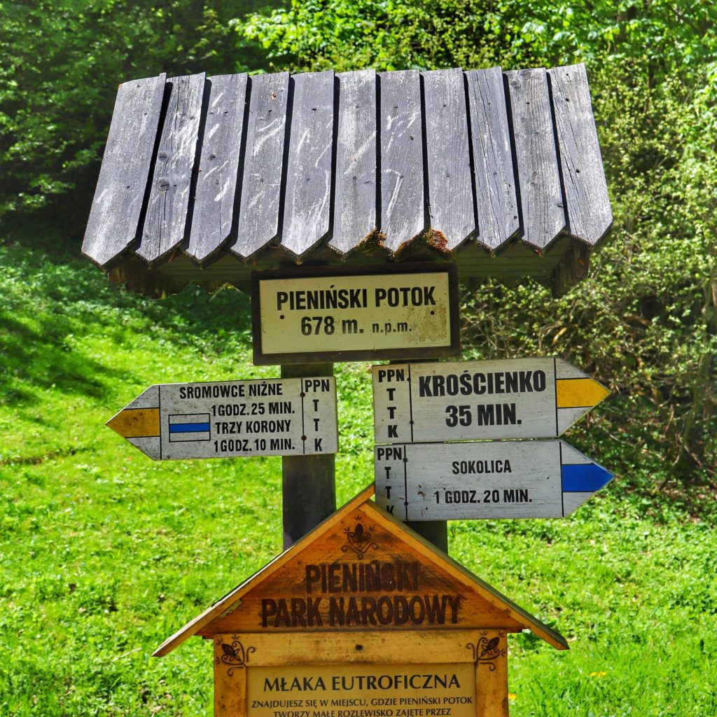 Biała tabliczka z napisem Pieniński Potok, drogowskazy, opis szlaku żółtego i niebieskie
