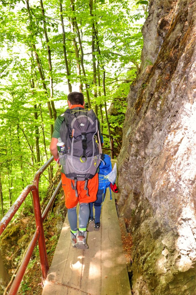 Turysta z dzieckiem przechodzący obok skalnej ściany drewnianym mostkiem
