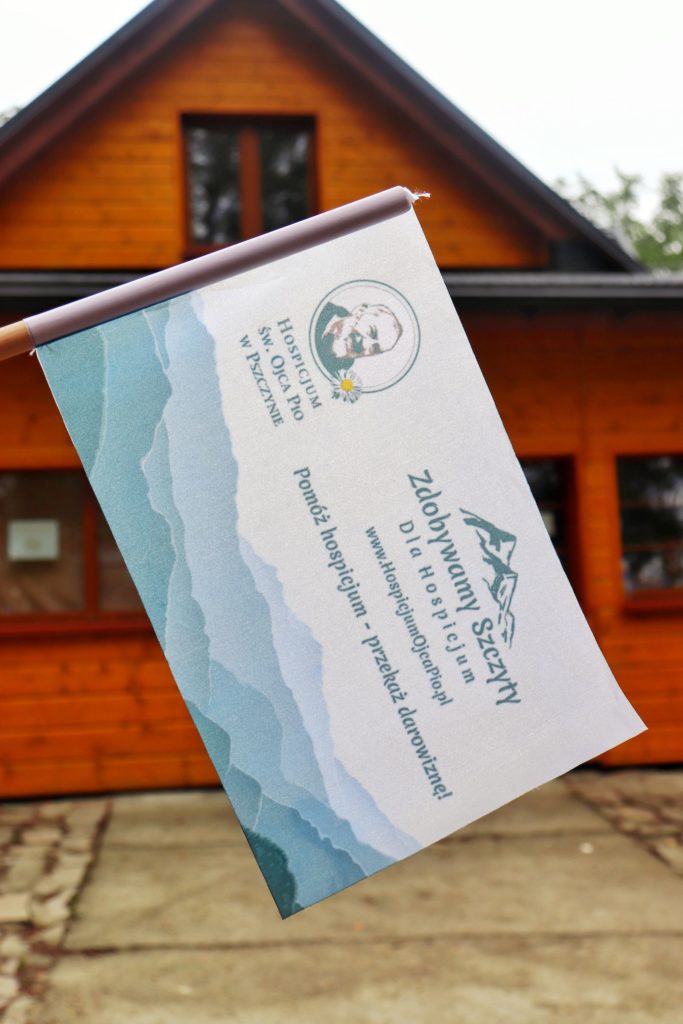 Flaga akcji zdobywamy szczyty dla hospicjum na tle budynku schroniska PTTK na Hrobaczej Łące