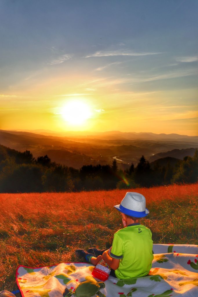 Mały turysta w kapeluszu siedzący na kocu piknikowym podziwiający zachód słońca z Polany Kretówki