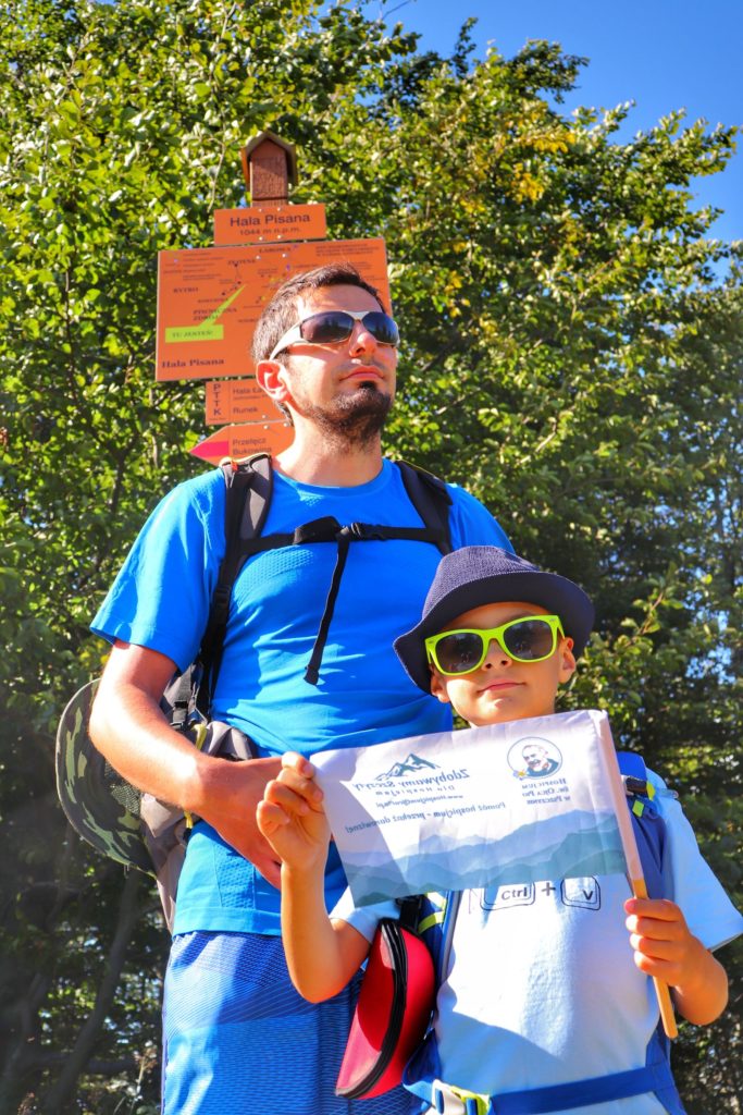 Turysta z dzieckiem w Beskidzie Sądeckim - Hala Pisana, pomarańczowa tablica, dziecko trzyma w rękach flagę akcji zdobywamy szczyty dla hospicjum