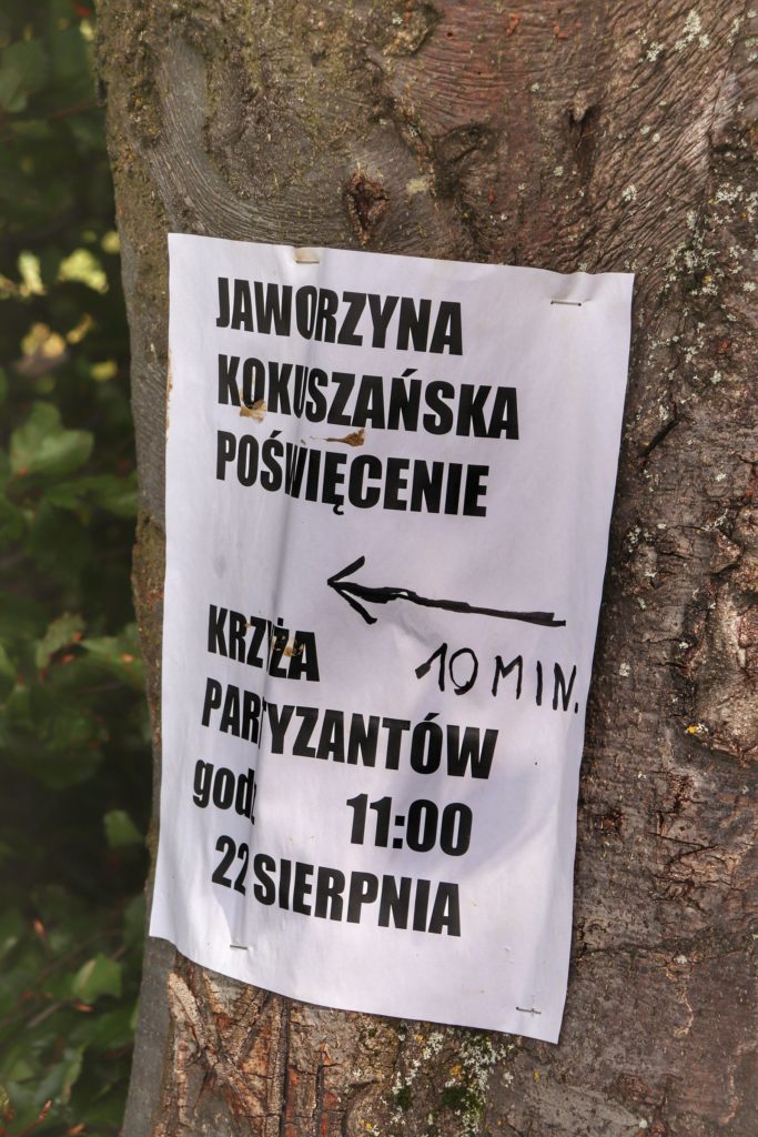 Biała kartka przyczepiona do drzewa informujące o poświęceniu Krzyża Partyzantów o godz. 11.00, 22 sierpnia