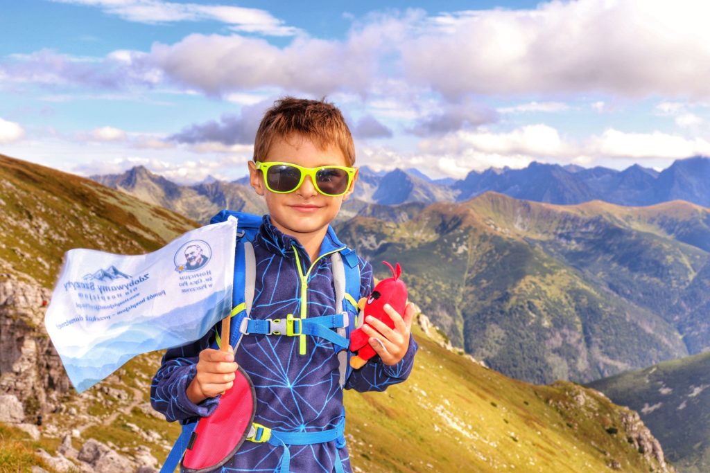 Zadowolone dziecko na Ciemniaku (Czerwone Wierchy), w tle tatrzańskie szczyty, chłopiec trzyma w ręce flagę akcji zdobywamy szczyty dla hospicjum