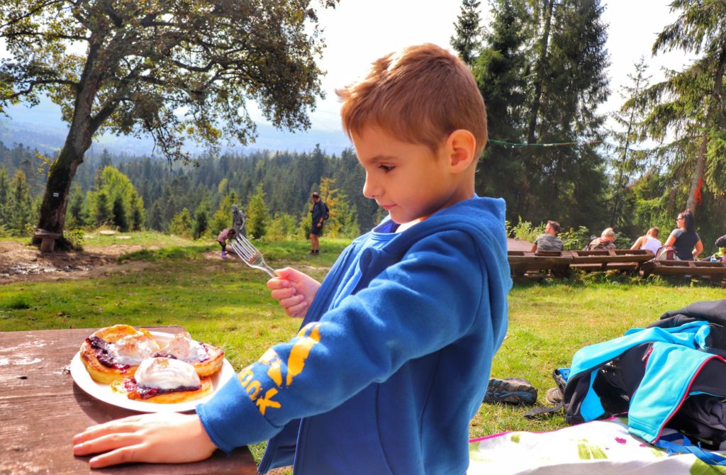 Dziecko jedzące racuchy ze śmietaną i borówkami zamówione w Kolibie na Łapsowej Polanie, polana przy schronisku, w tle inni turyści