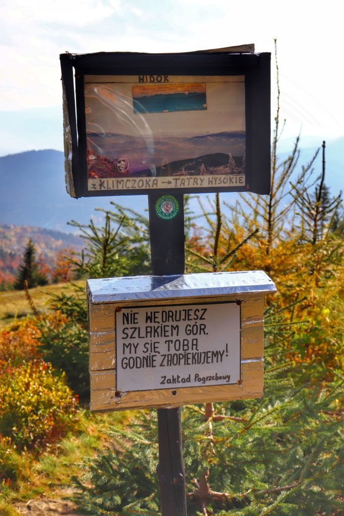 Tabliczki na szczycie Klimczok w Beskidzie Śląskim, panorama na Tatry Wysokie oraz tabliczka z napisem - Nie wędrujesz szlakiem gór. My się Tobą godnie zaopiekujemy. Zakład Pogrzebowy