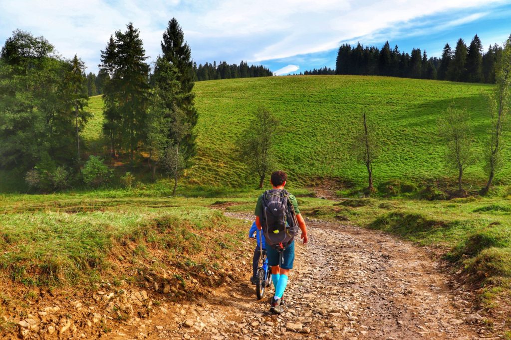 Turysta prowadzący dziecko na rowerku, szlak czerwony na Żar, wieś Dursztyn, szeroka kamienista droga idąca między polami