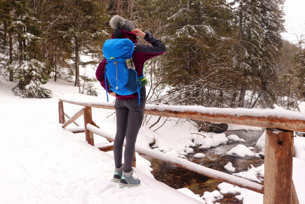 Turystka stojąca na drewnianym, zasypanym przez śnieg moście, podziwiająca potok przepływający niopodal miejsca noszącego nazwę - Rozdroże Salviovy Prameń w Tatrach Słowackich