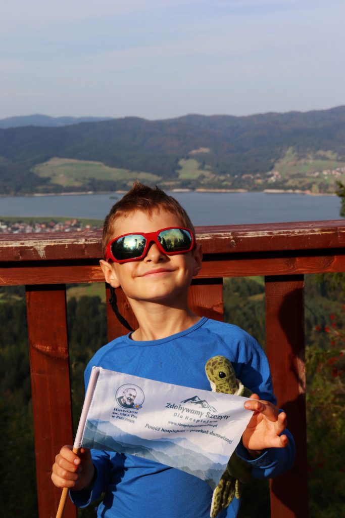Uśmiechnięte dziecko trzymające w rękach flagę akcji zdobywamy szczyty dla hospicjum, stojące na drewnianej wieży widokowej Żar w Pieninach, w tle widoczne Jezioro Czorsztyńskie