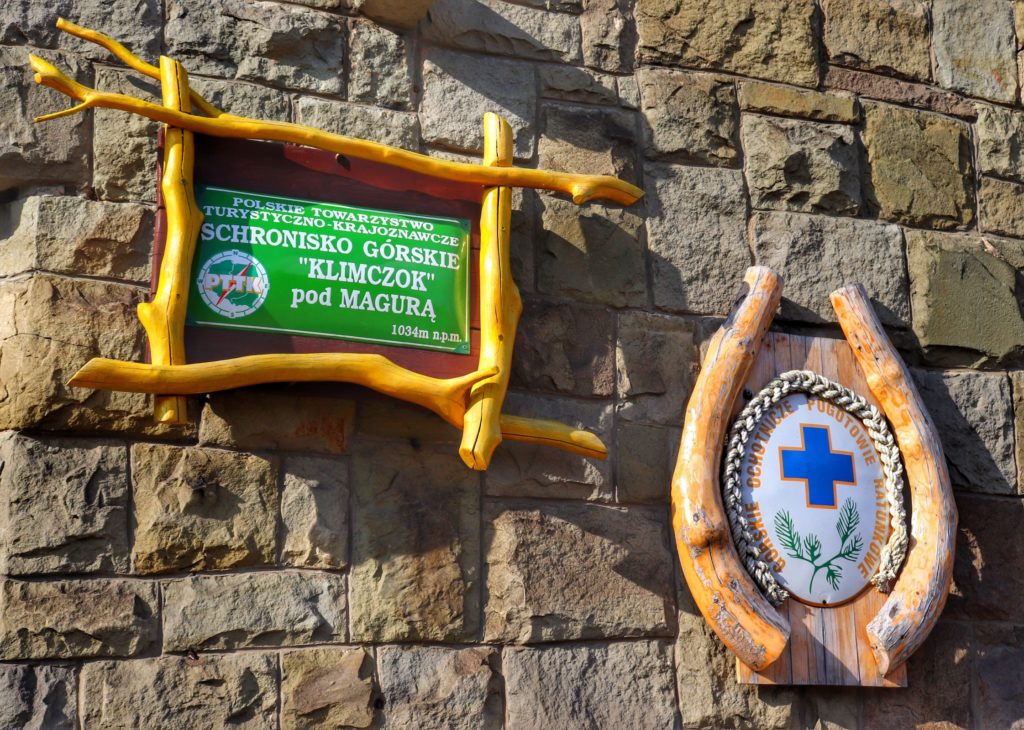 Zielona tablica wisząca na budynku schroniska z napisem - SCHRONISKO GÓRSKIE KLIMCZOK POD MAGURĄ, znaczek górskiego ochotniczego pogotowia ratunkowego