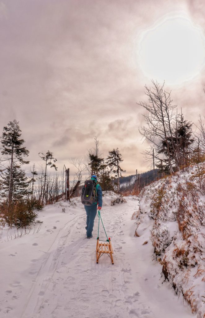 Mężczyzna ciągnący drewniane sani, idący zaśnieżoną ścieżką w Tatrach Słowackich, na niebie słońce, gęste chmury