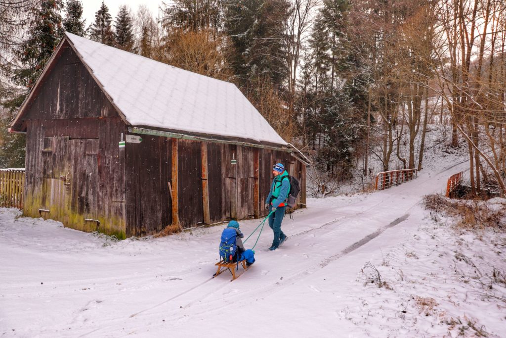 Mężczyzna ciągnący dziecko siedzące na drewnianych sankach. ulica Danielki - Ujsoły - początek szlaku zielonego na szczyt Muńcuł