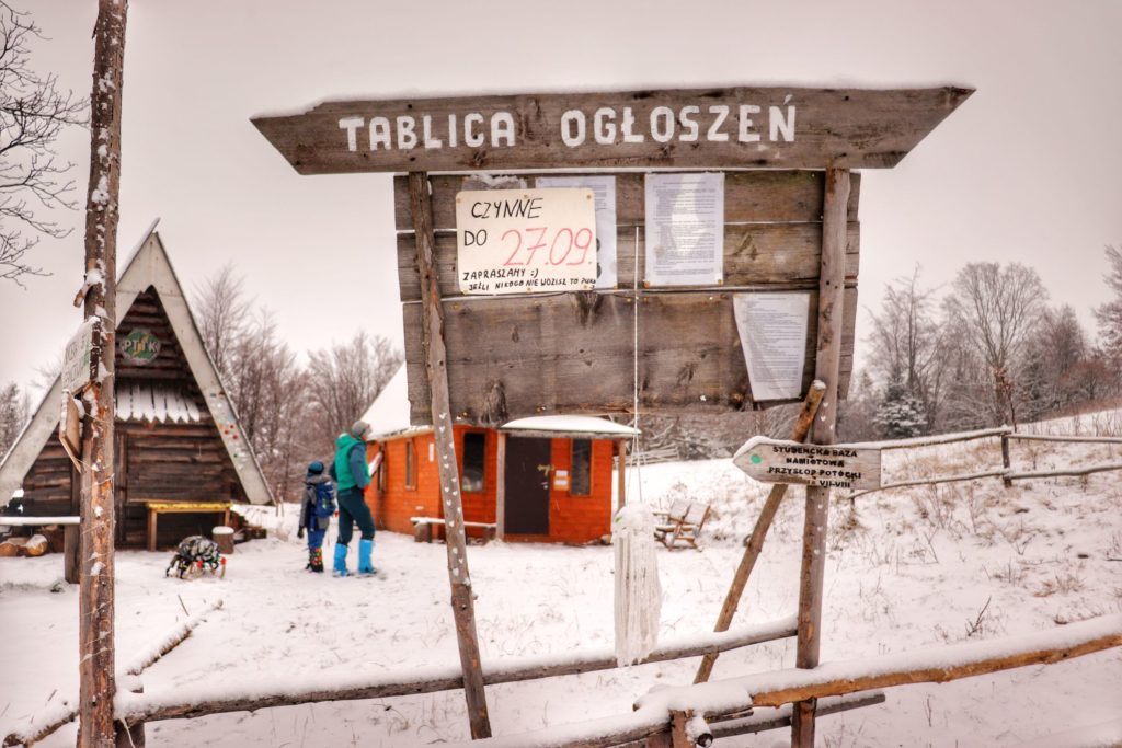 Tablica ogłoszeń przy studenckiej bazie namiotowej Przysłop Potócki, zima, w tle turysta z dzieckiem oraz drewniane chatki na bazie namiotowej