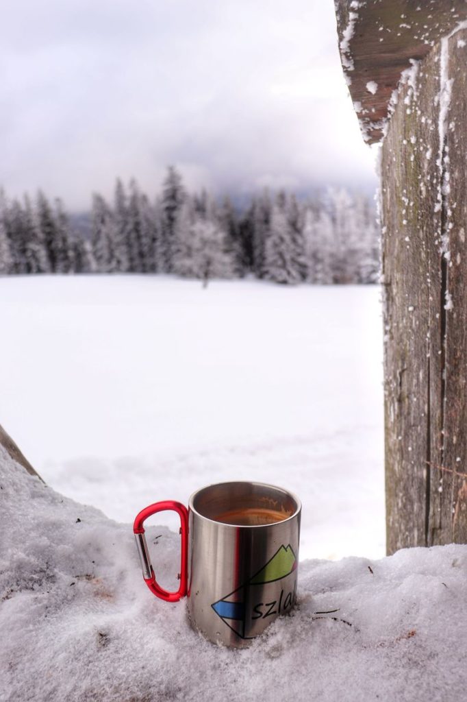 Metalowy kubek z kawą leżący w śniegu, w tle zaśnieżone drzewa