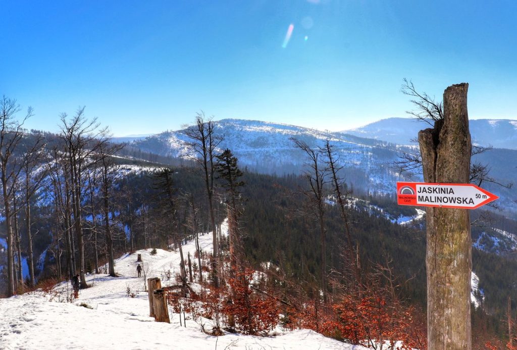 Drewniany słup z czerwono - białą tabliczką z napisem Jaskinia Malinowska, śnieg, krajobraz górski