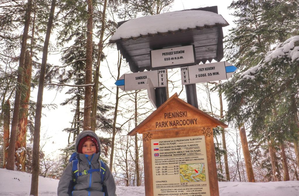 Dziecko siedzące na betonowym słupku przy tablicy informacyjnej Pienińskiego Parku Narodowego zimą na Przełęczy Sosnów