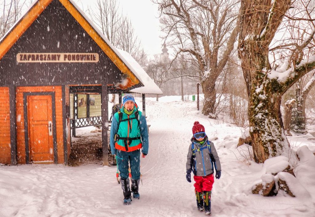 Młody mężczyzna z sześcioletnim dzieckiem wchodzący do Wąwozu Homole w Pieninach, mroźny, zimowy dzień, sypiący śnieg, w tle drewniana budka, w której jest kasa pienińskiego parku narodowego