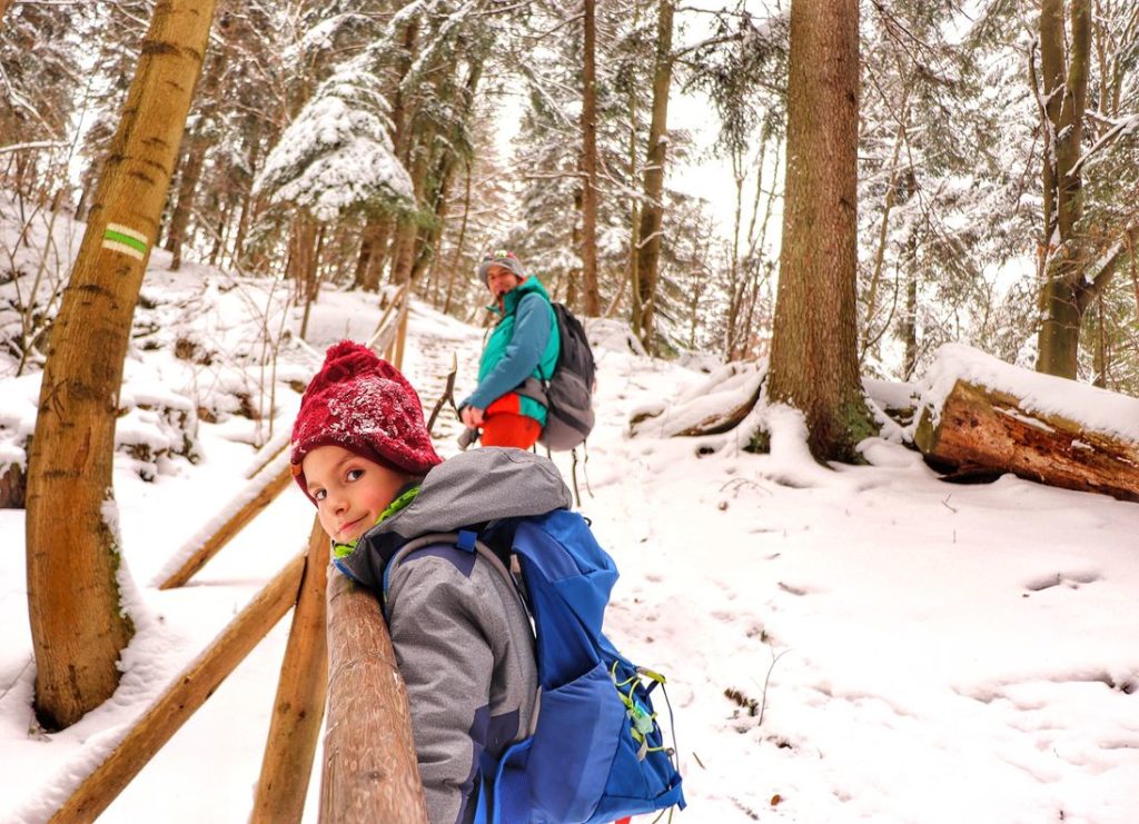 Zadowolone dziecko opierające głowę o drewnianą barierkę na zielonym szlaku na Sokolicę z Krościenka, w tle tata dziecka, drewniane schody na szlaku, las, zima