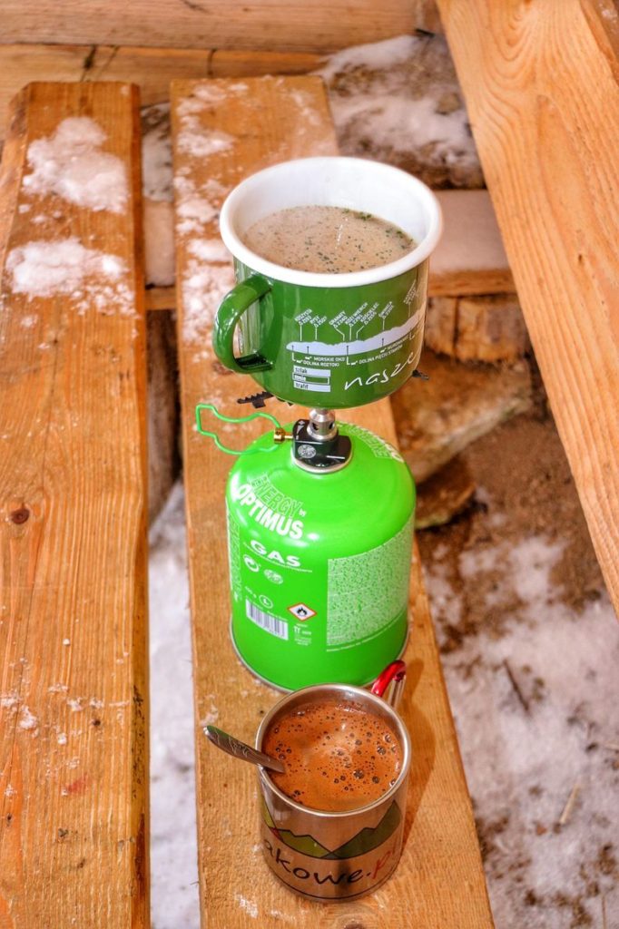 Zielona butla gazowa, na której położony jest kubek z zupą, obok na drewnianej ławie leży metalowy kubek z gorącą kawą