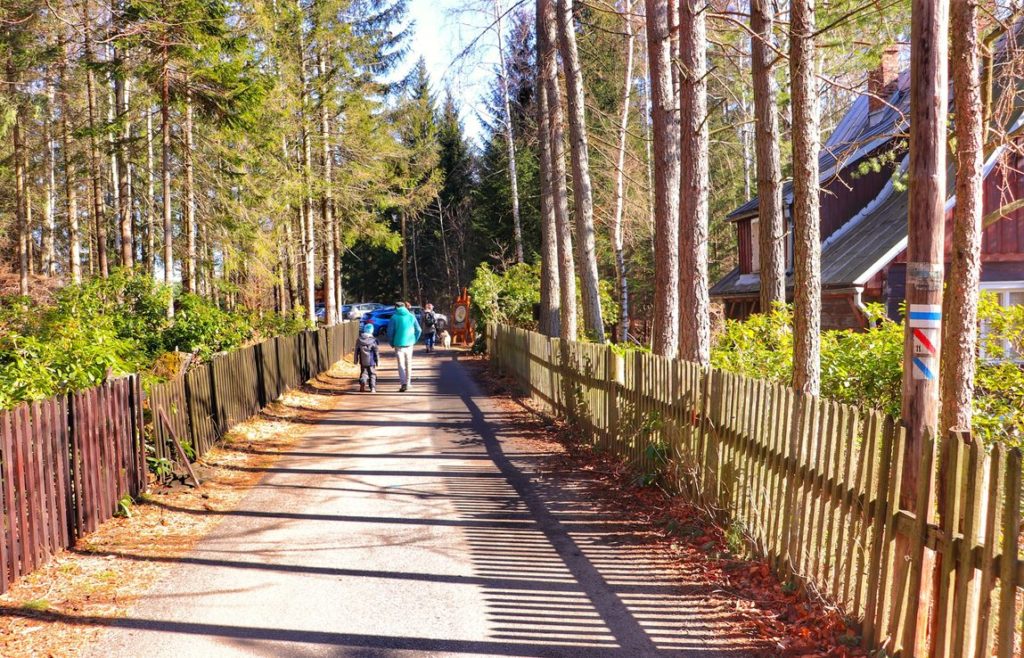 Asfaltowa droga na niebieskim szlaku w Szklarskiej Porębie prowadząca obok znajdującego się po prawej stronie ulicy muzeum - domu Wlastimila Hofmana, płot, w oddali turyści