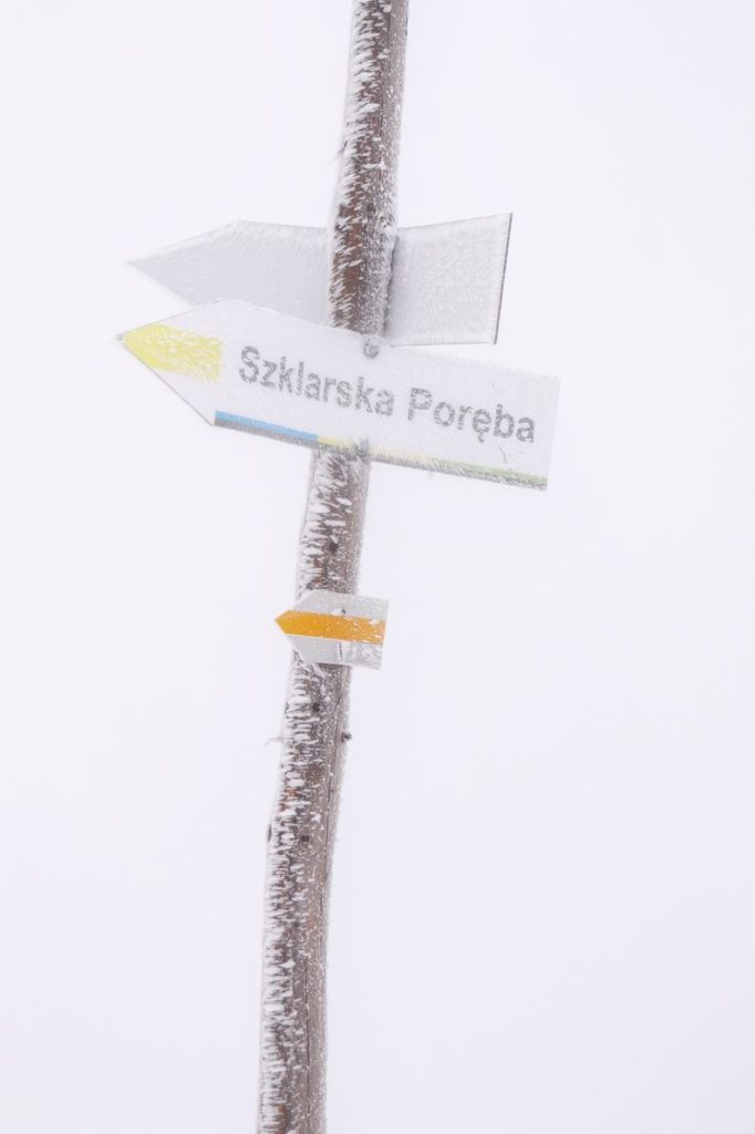 Biała tabliczka w kształcie strzałki wskazująca kierunek do Szklarskiej Poręby (szlak żółty)