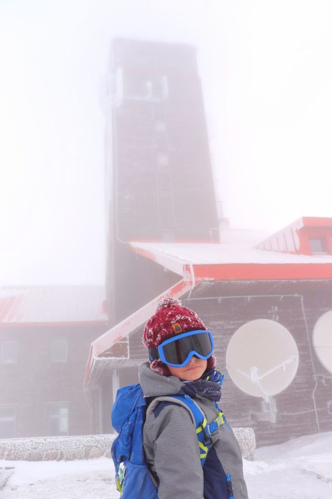 Dziecko w goglach na tle budynku znajdującego się na Śnieżnych Kotłach w Karkonoszach, mgła, zimowy dzień