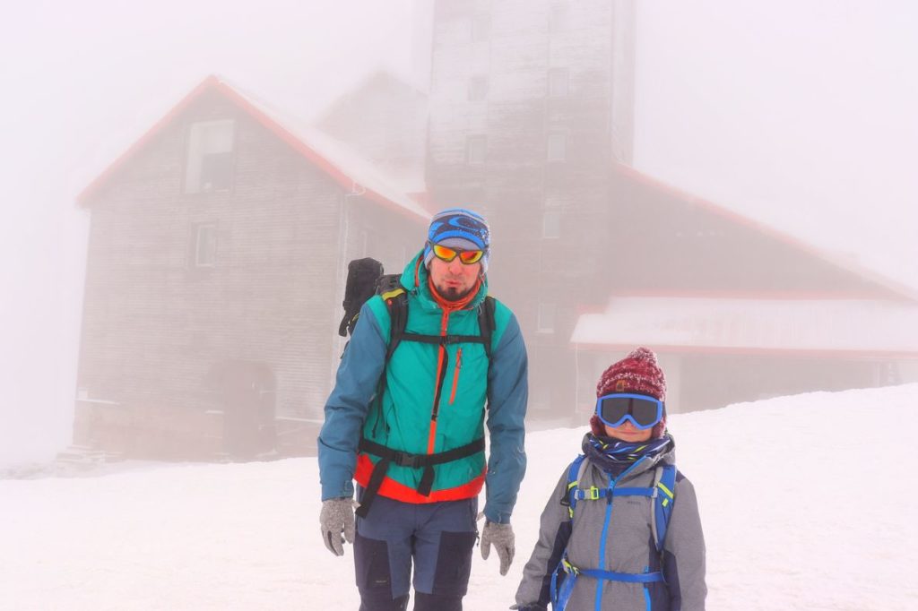 Turysta z dzieckiem, zimowe warunki, mgła, w tle budynek, w którym znajduje się Radiowo – telewizyjny Ośrodek Nadawczy Śnieżne Kotły