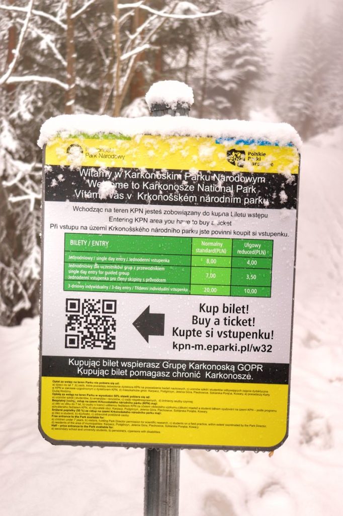 Żółto biała tablica zasypana przez śnieg z cennikiem biletów w Karkonoskim Parku Narodowym