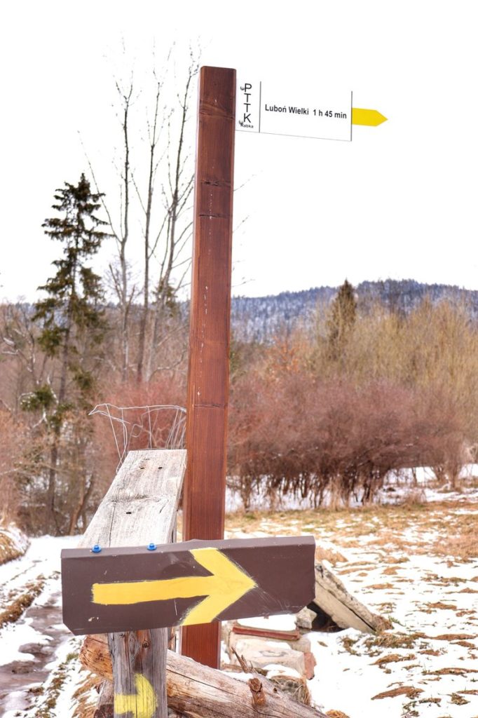 Biała tabliczka wskazująca kierunek przebiegu żółtego szlaku na Luboń Wielki (skręt w prawo), czas przejścia 1 h 45 min