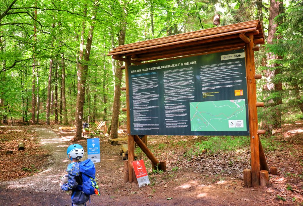 Las, dziecko w kasku rowerowym, duża drewniana tablica opisująca regulamin trasy rowerowej CHEŁMSKA TRAILS w Koszalinie
